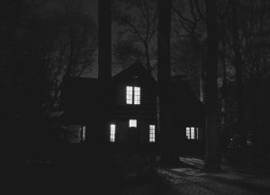 A haunting black and white image of a house surrounded by trees at night, showcasing an eerie atmosphere.