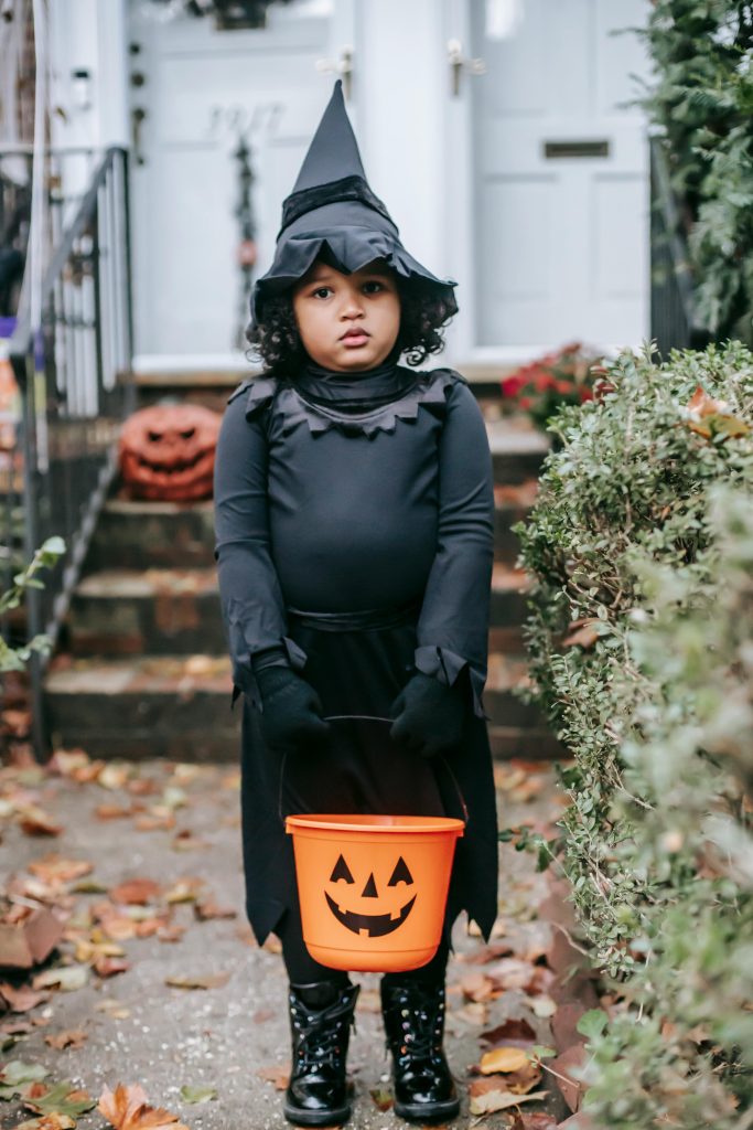 Full body little ethnic girl wearing witch costume standing with Trick or Treat bucket near cottage entrance and looking at camera
